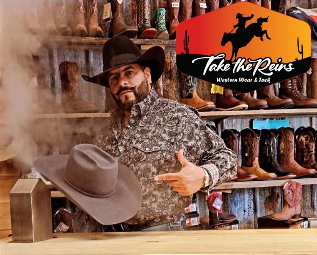 Tex Ruiz holding a felt cowboy hat in front of the boot wall at Take the Reins